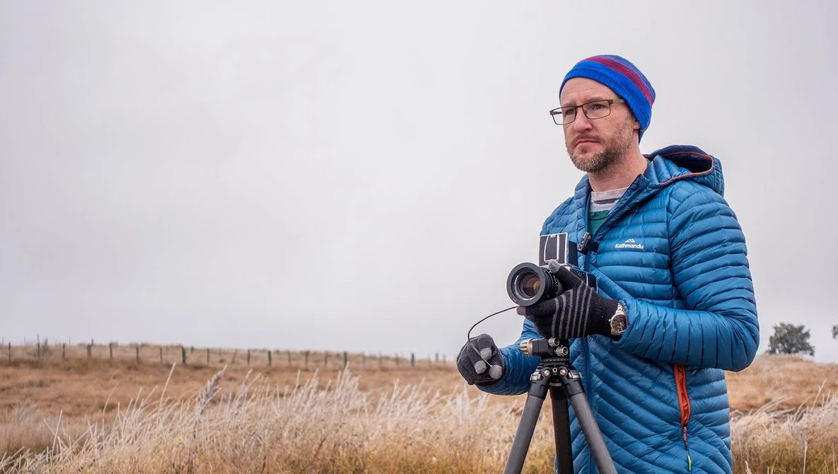 Filmmaker in blue jacket and beanie operating a cinema camera on tripod in an open field.