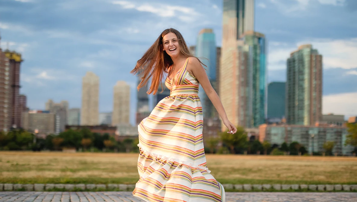 Woman in striped sundress poses on waterfront promenade with urban skyline behind her.