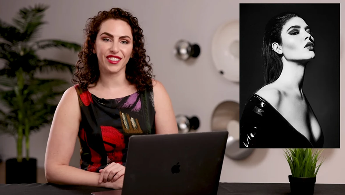 Woman with curled auburn hair sitting at desk with laptop, wearing colorful patterned top with red lipstick. Inset black and white portrait of woman in profile.