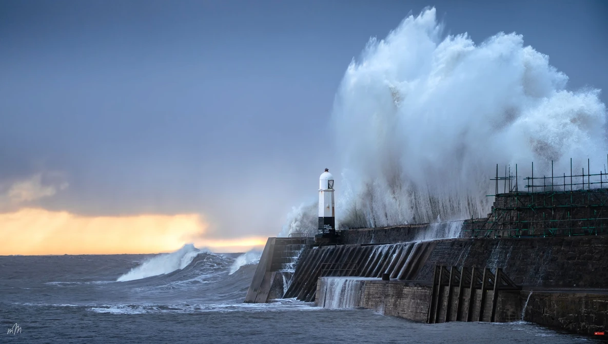 Massive wave crashes against a lighthouse and harbor breakwater during stormy conditions.