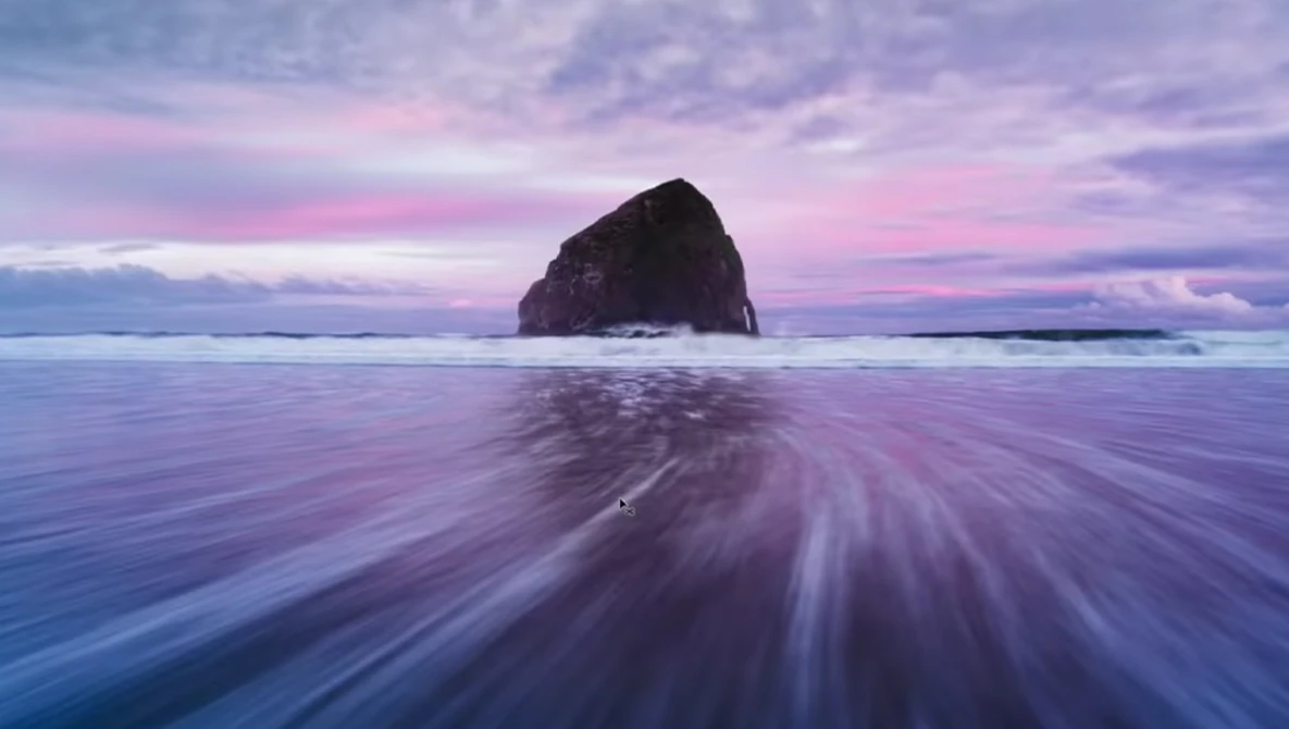 A dark rocky sea stack rises from the ocean during twilight, with smooth water motion blur in the foreground.