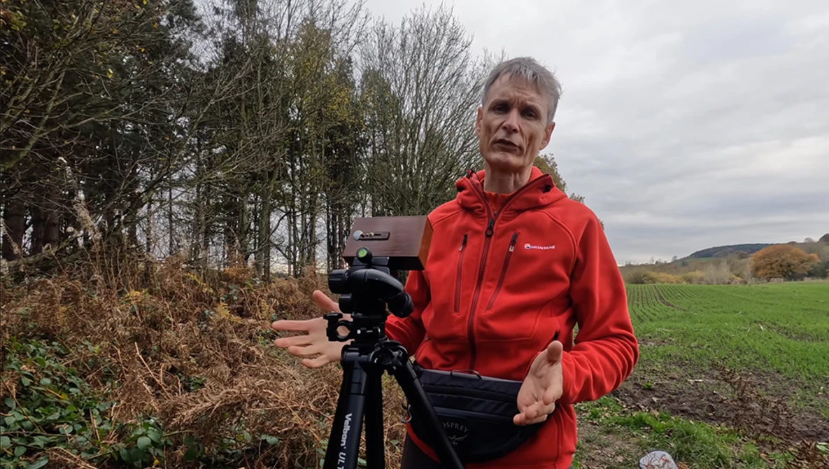Videographer in red jacket holding cinema camera on tripod in rural landscape.