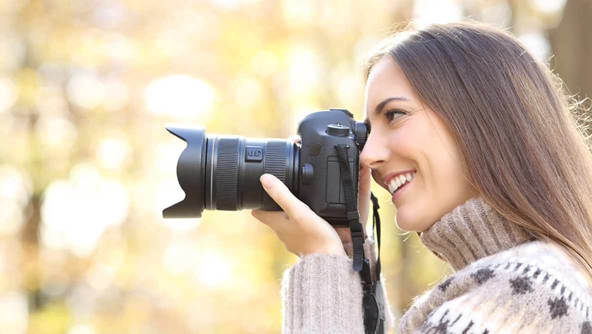 Woman holding DSLR camera with telephoto lens while smiling at camera outdoors.