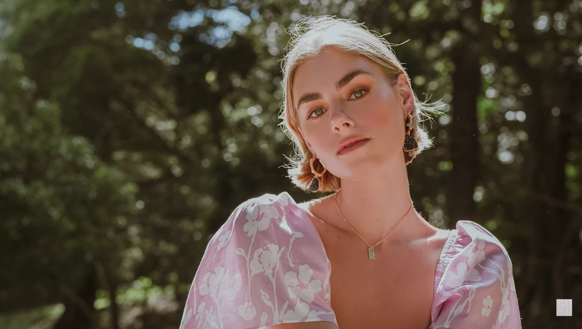 Portrait of a woman with platinum blonde hair wearing a pink patterned blouse against a blurred forest backdrop.
