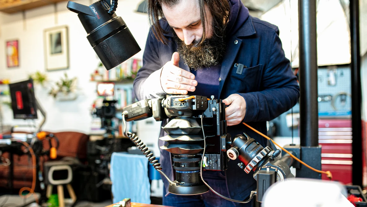 Photographer wearing navy jacket examines camera lens components at a workbench in a studio setting.