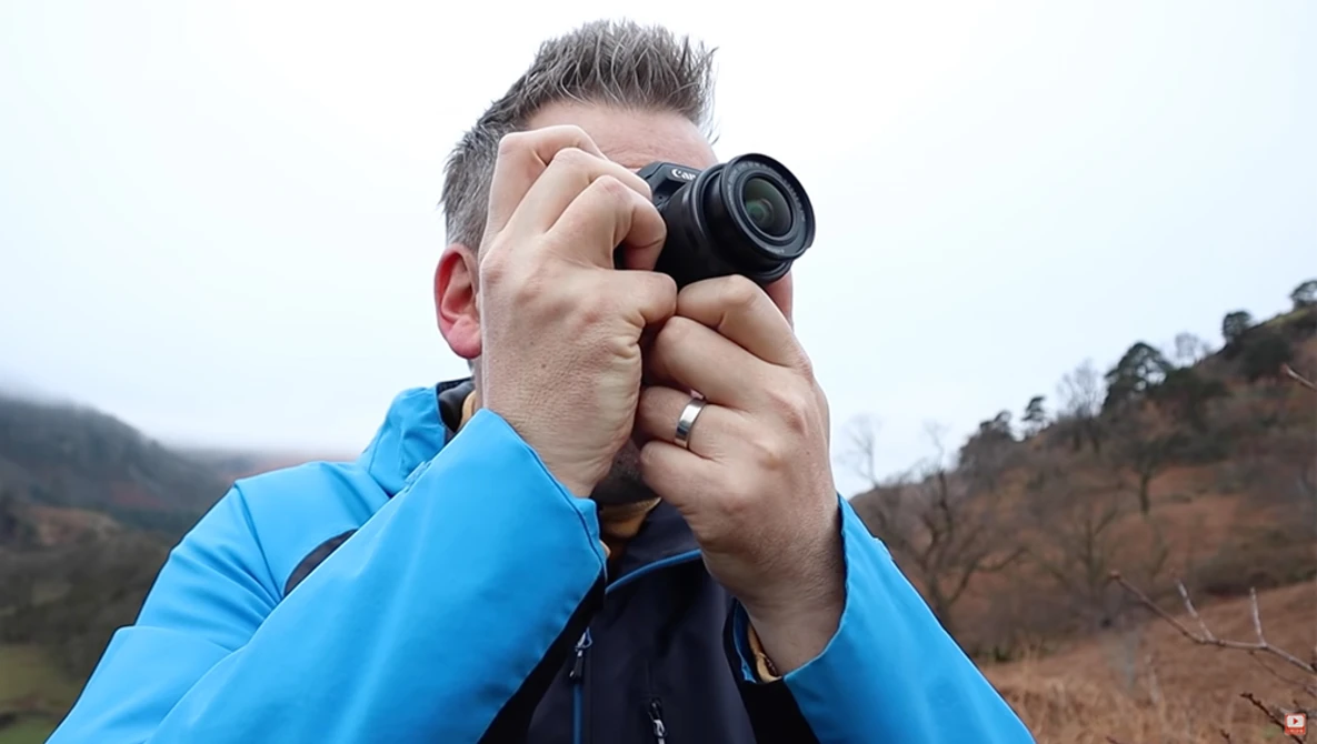 Photographer holding a compact camera to his eye, wearing a blue jacket in an outdoor hillside setting.
