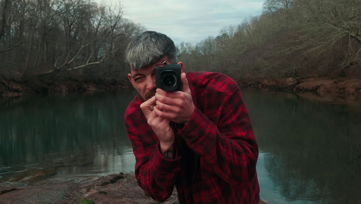 Photographer in red plaid shirt crouching by a river, aiming a telephoto lens toward the camera.