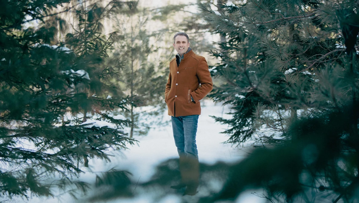 Man in rust-colored jacket standing among evergreen trees in winter landscape.