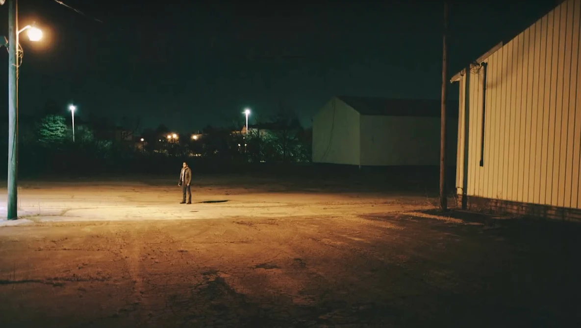 Solitary figure standing in an empty lit parking area at night beside an industrial metal building.