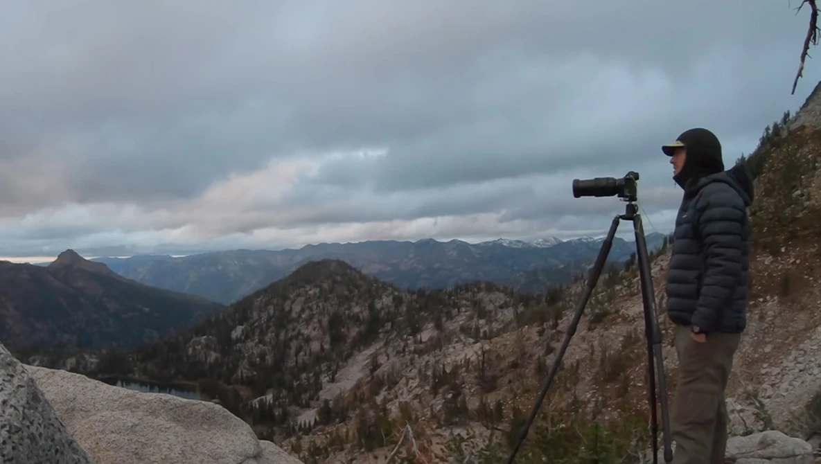 Photographer with telescope observing distant mountain landscape from high elevation.
