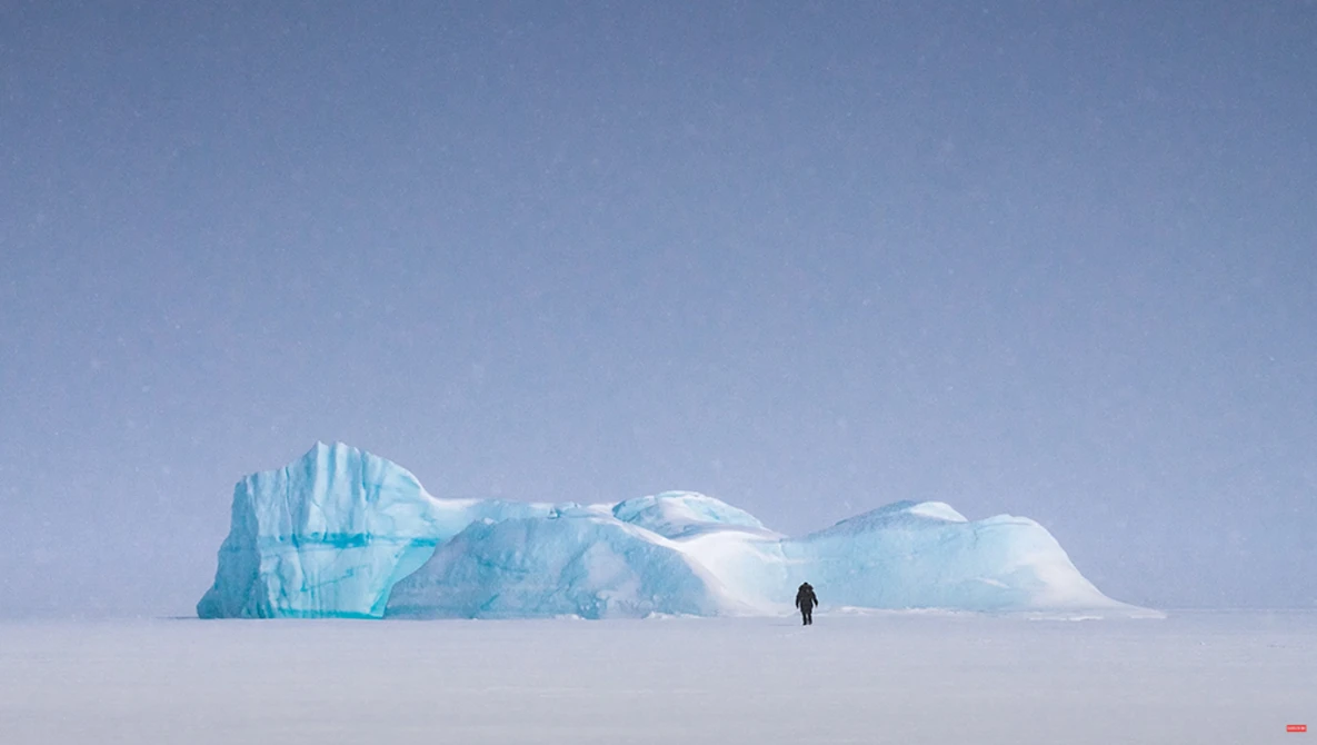 Solitary figure standing on snow facing a large blue iceberg under clear sky.