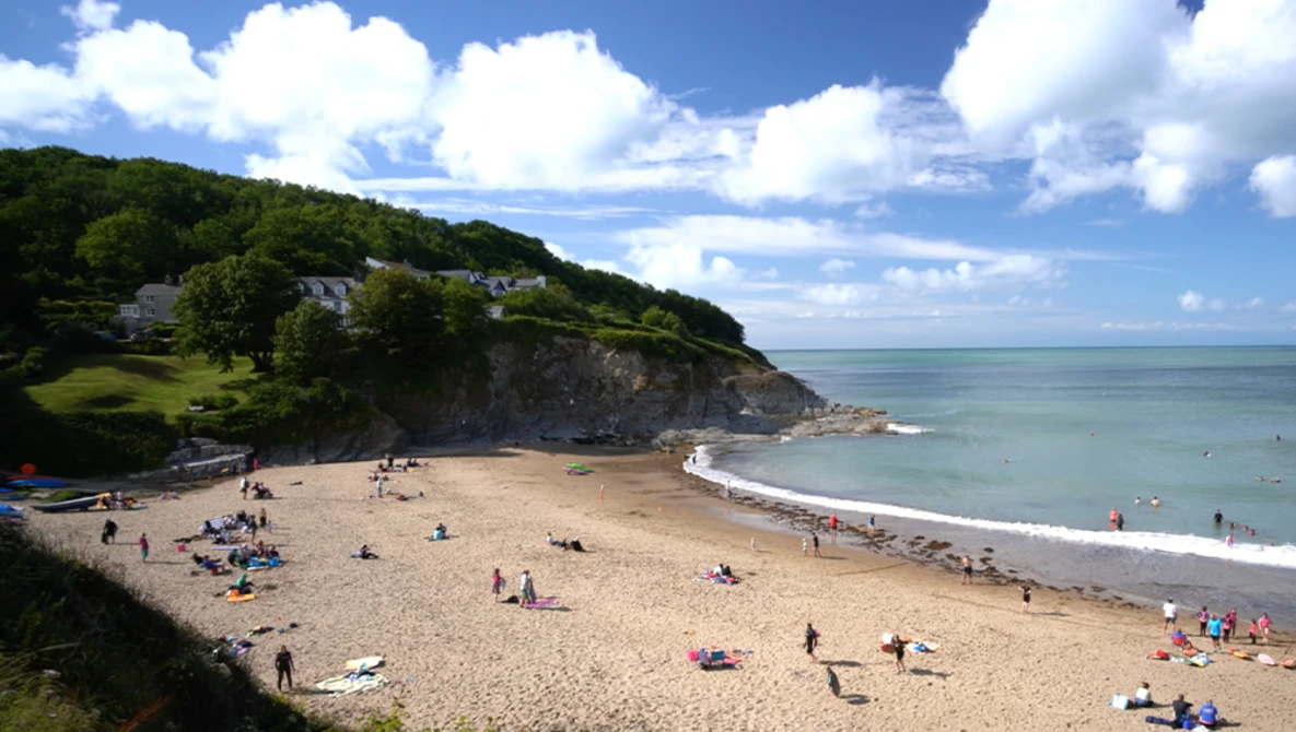 Scenic coastal beach with sandy shores, beachgoers, and dramatic green cliffs under blue sky with white clouds.