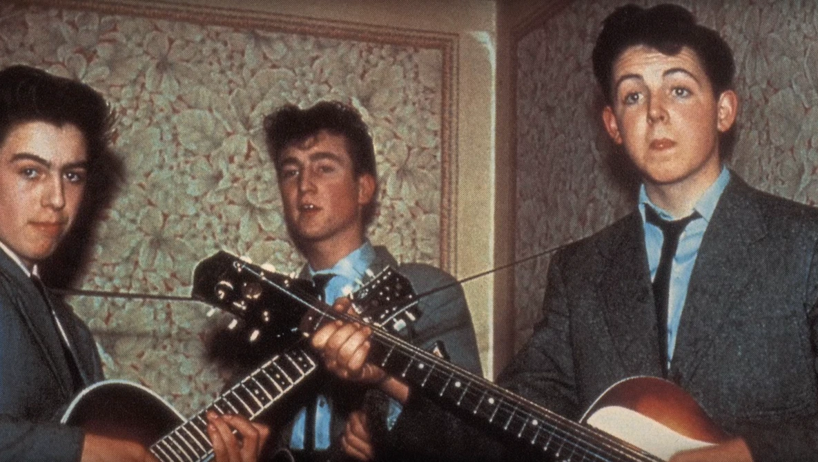 Three young men with acoustic guitars posed against floral wallpaper in an indoor setting.