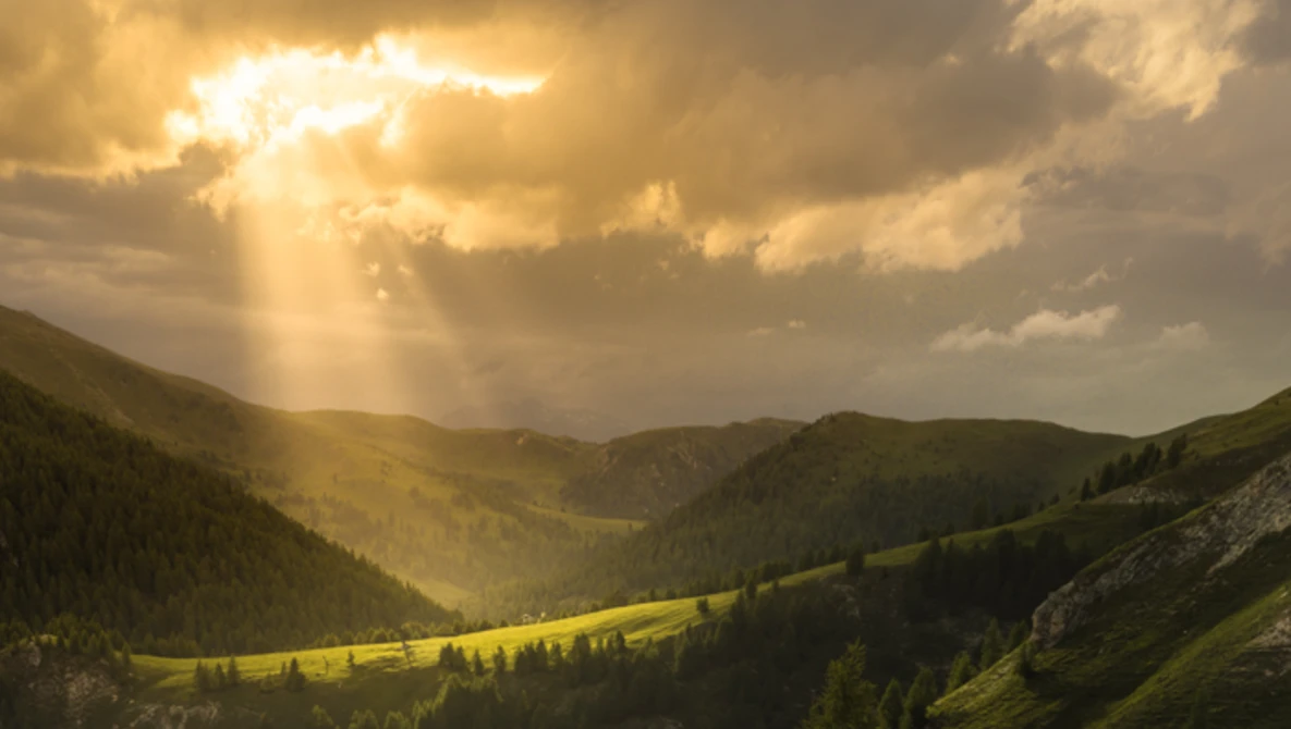 Mountain valley with golden sunlight breaking through clouds, forested slopes, and meadow in afternoon light.