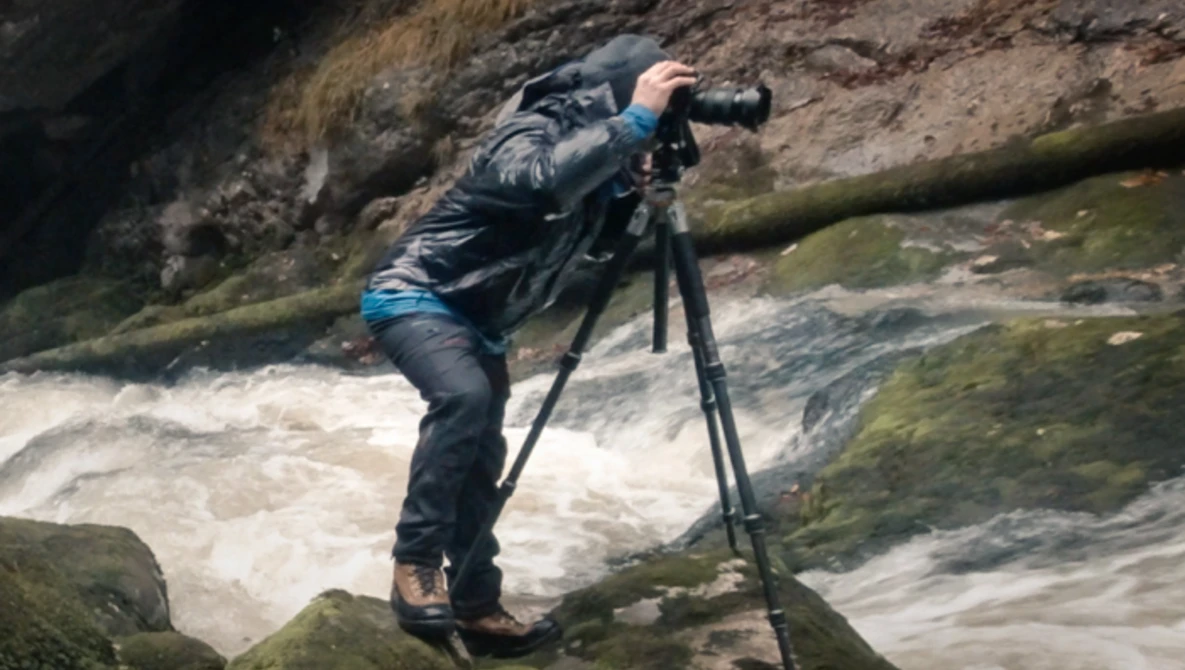 Photographer on tripod shooting whitewater rapids flowing over moss-covered rocks.