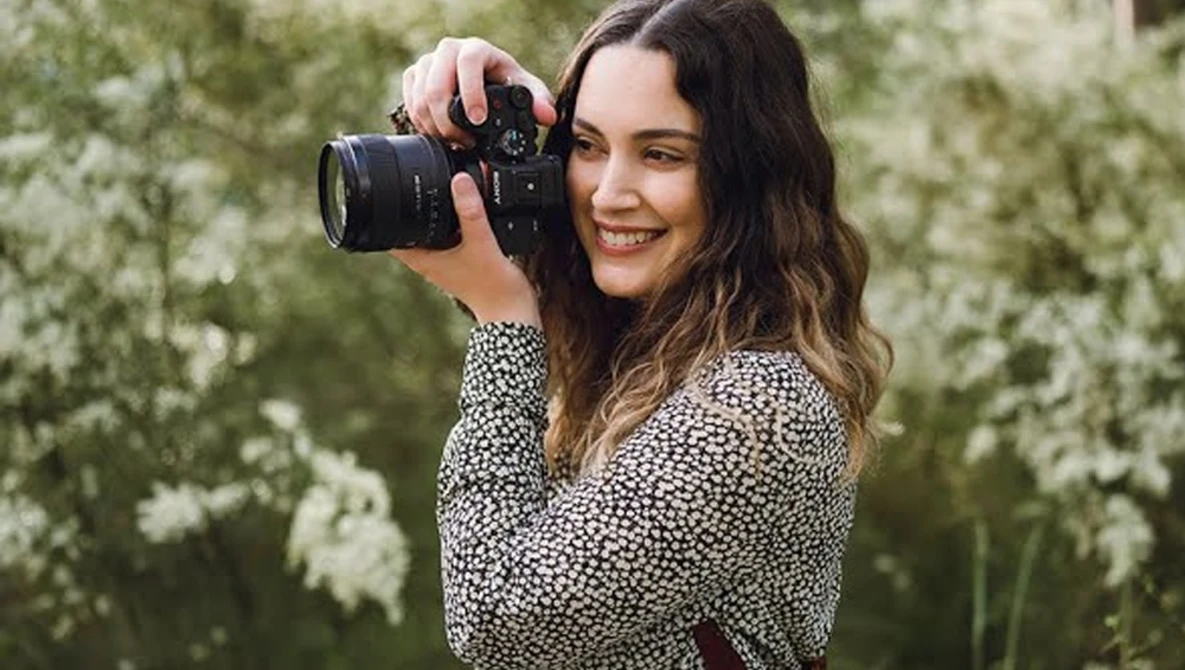 Woman holding a professional camera with telephoto lens, smiling at camera against blooming white flowers.