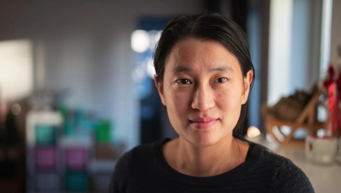 Portrait of a woman with shoulder-length dark hair wearing a black t-shirt, looking directly at the camera in what appears to be a creative workspace.