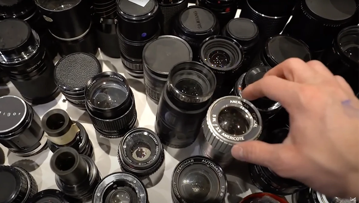 Hand selecting a vintage manual camera lens from a collection of black film lenses arranged on a white surface.