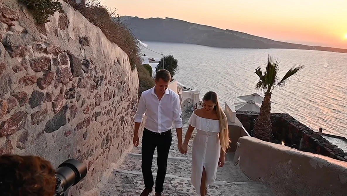 Couple walking hand-in-hand along a coastal stone wall at golden hour sunset.