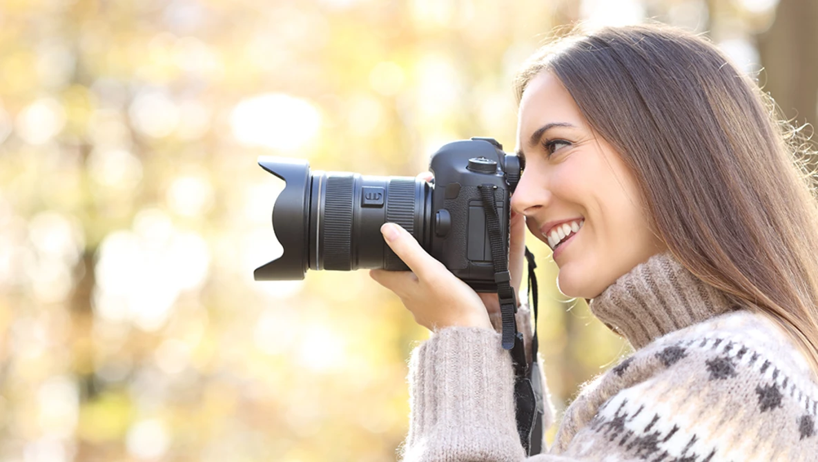 Woman holding a DSLR camera to her face while smiling outdoors.