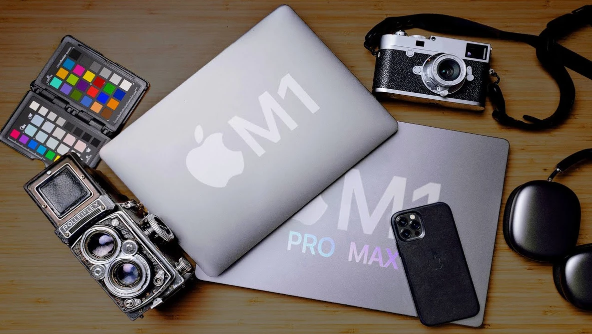 Apple M1 and M2 Pro Max laptops displayed with photography gear including a color checker chart, vintage film camera, rangefinder camera, smartphone, and over-ear headphones on wooden surface.