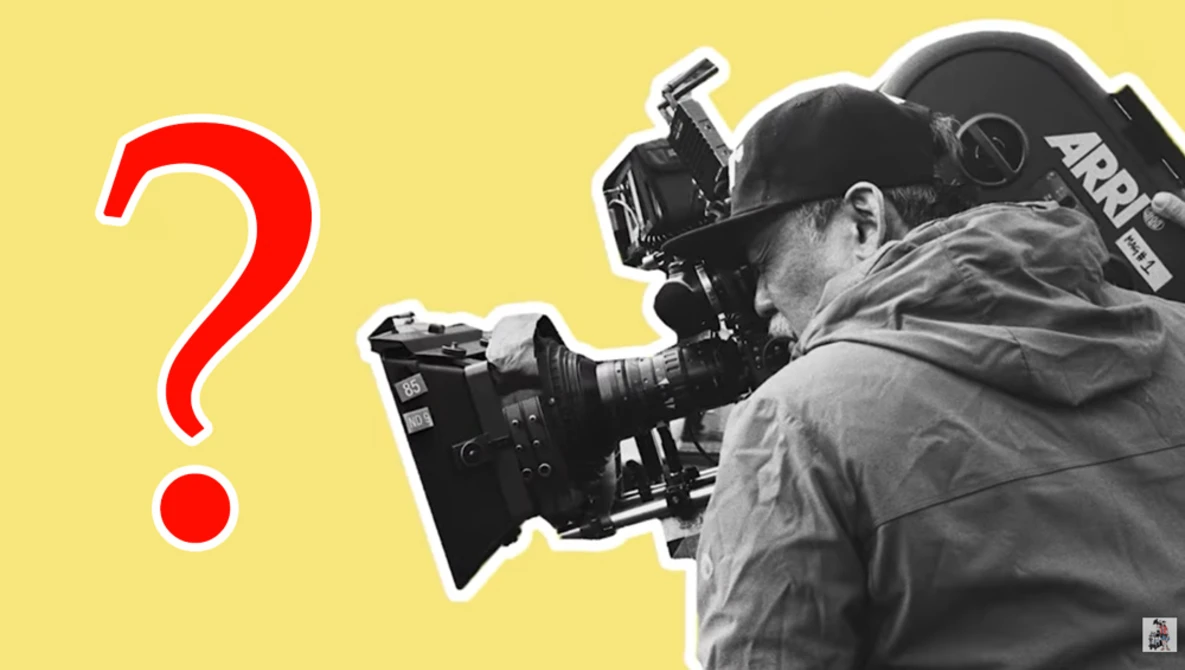 Cinematographer operating a professional film camera while wearing winter clothing against a yellow background.