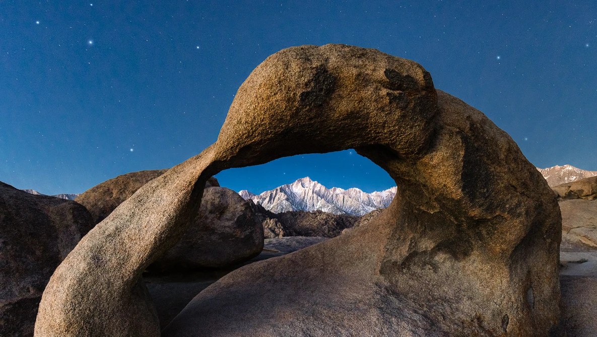 Granite boulder arch frames snow-capped mountain peak under starry night sky.