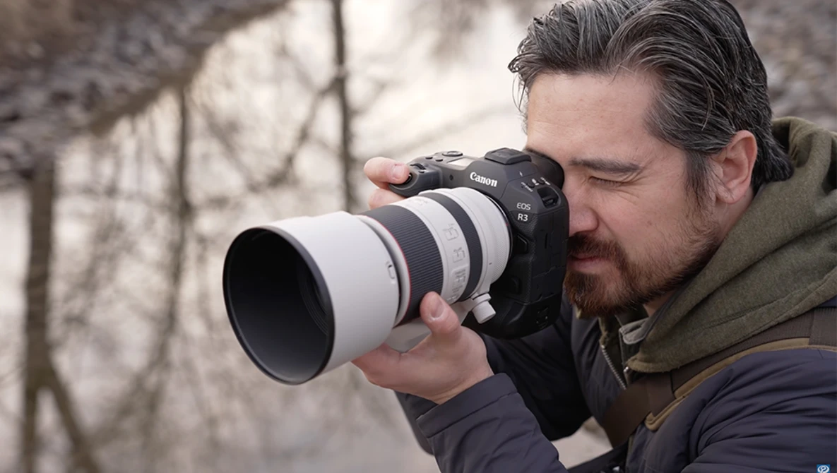Photographer holding a DSLR camera with a white telephoto lens, composing a shot outdoors.