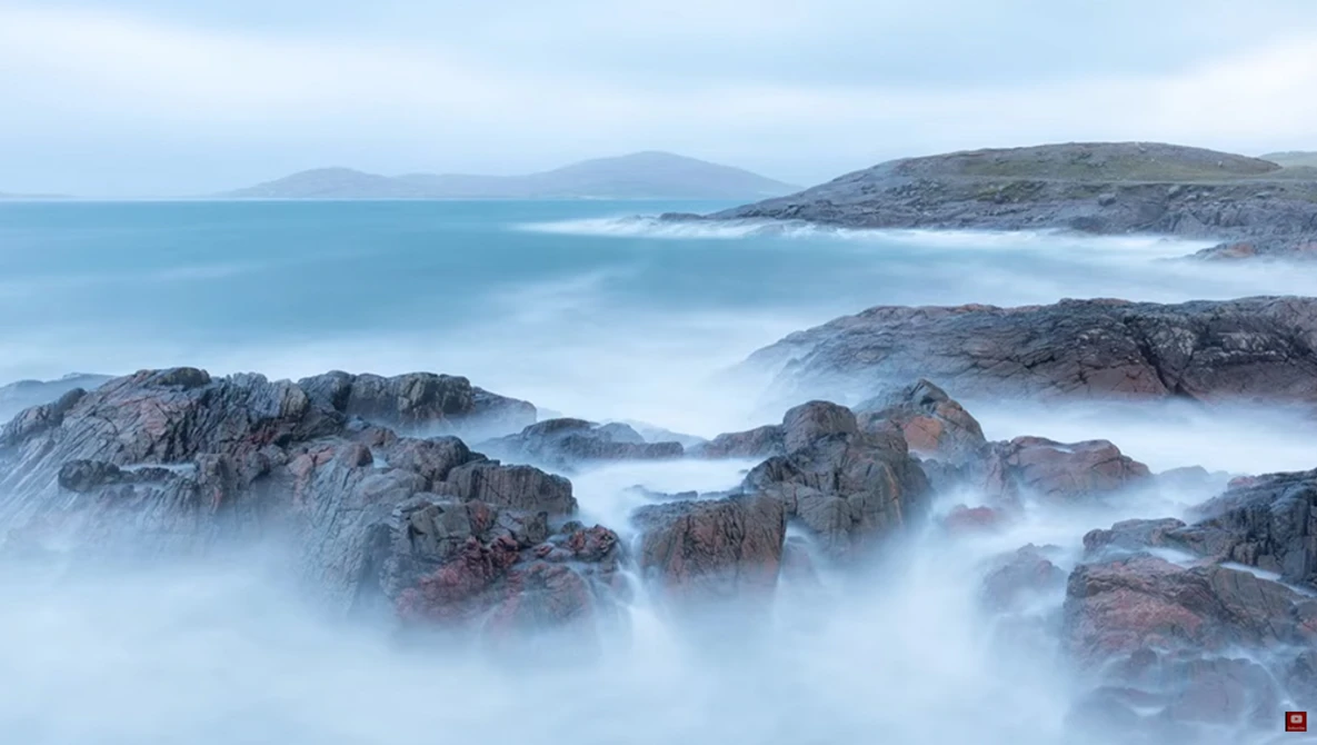 Rocky coastal rocks enveloped in misty seawater with mountains visible across a calm bay.
