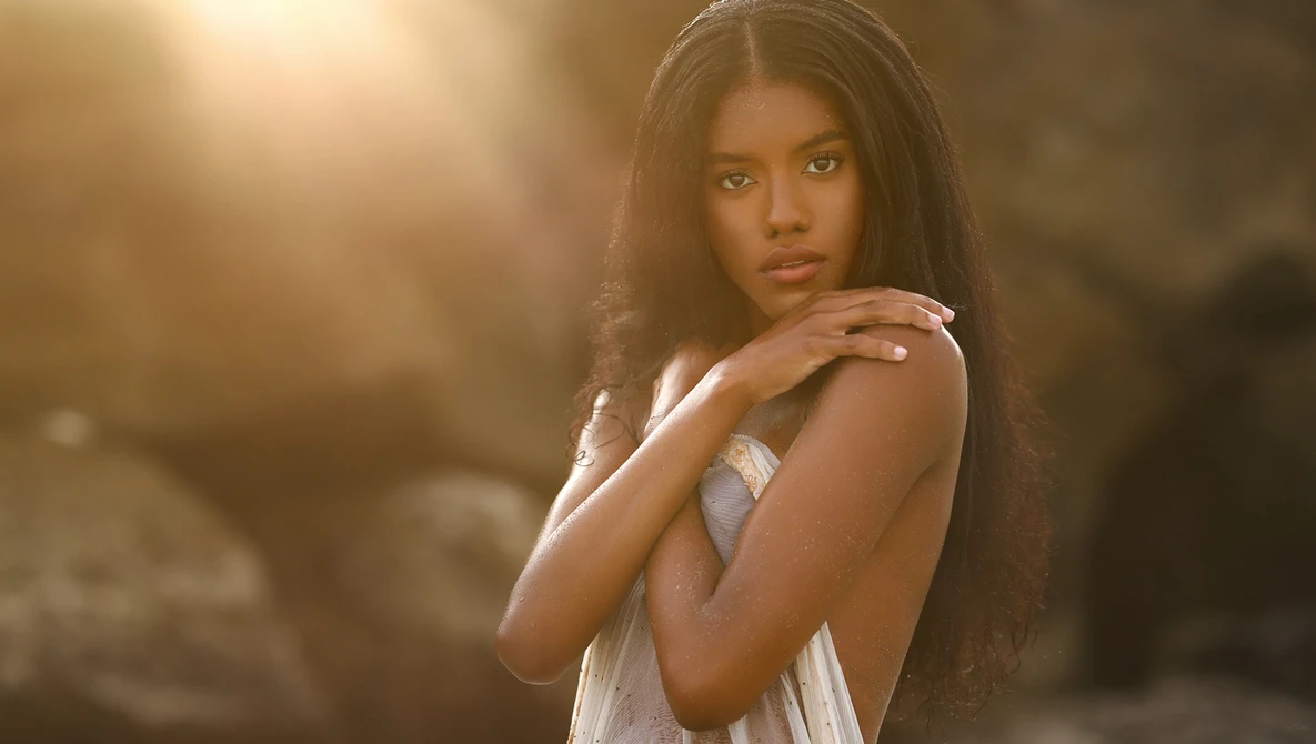 Woman with long dark hair wearing a white sleeveless top, posed against a warm, backlit background.