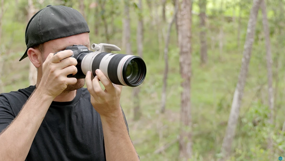 Photographer holding a telephoto lens camera to his face in a forest setting.