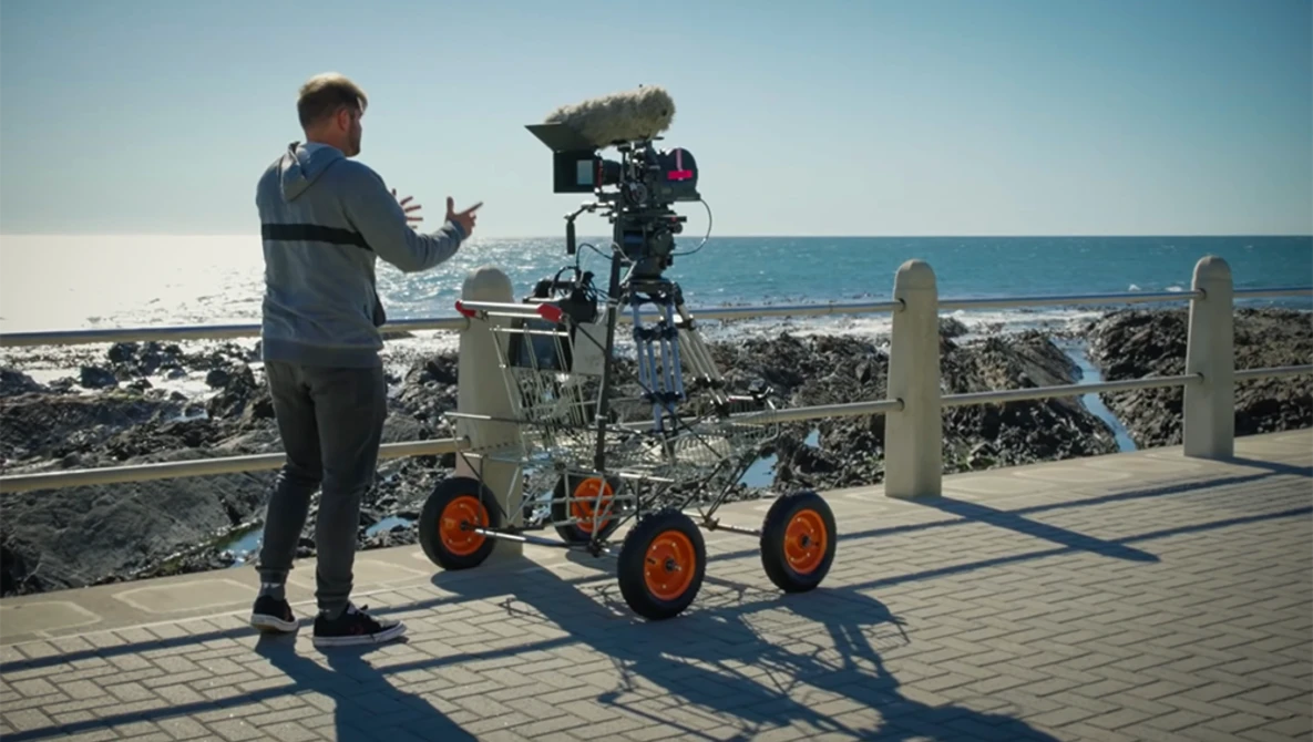 Filmmaker gestures while operating a camera rig mounted on a wheeled dolly at a coastal location.