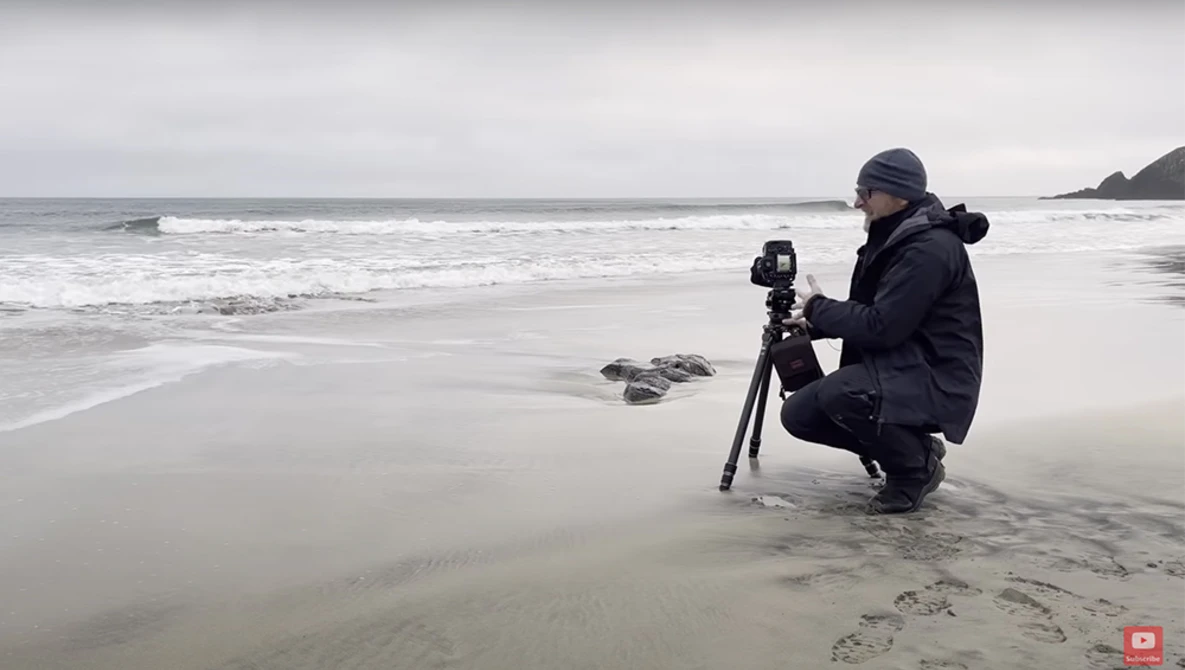 Photographer crouching on a beach with a tripod-mounted camera while framing ocean waves.