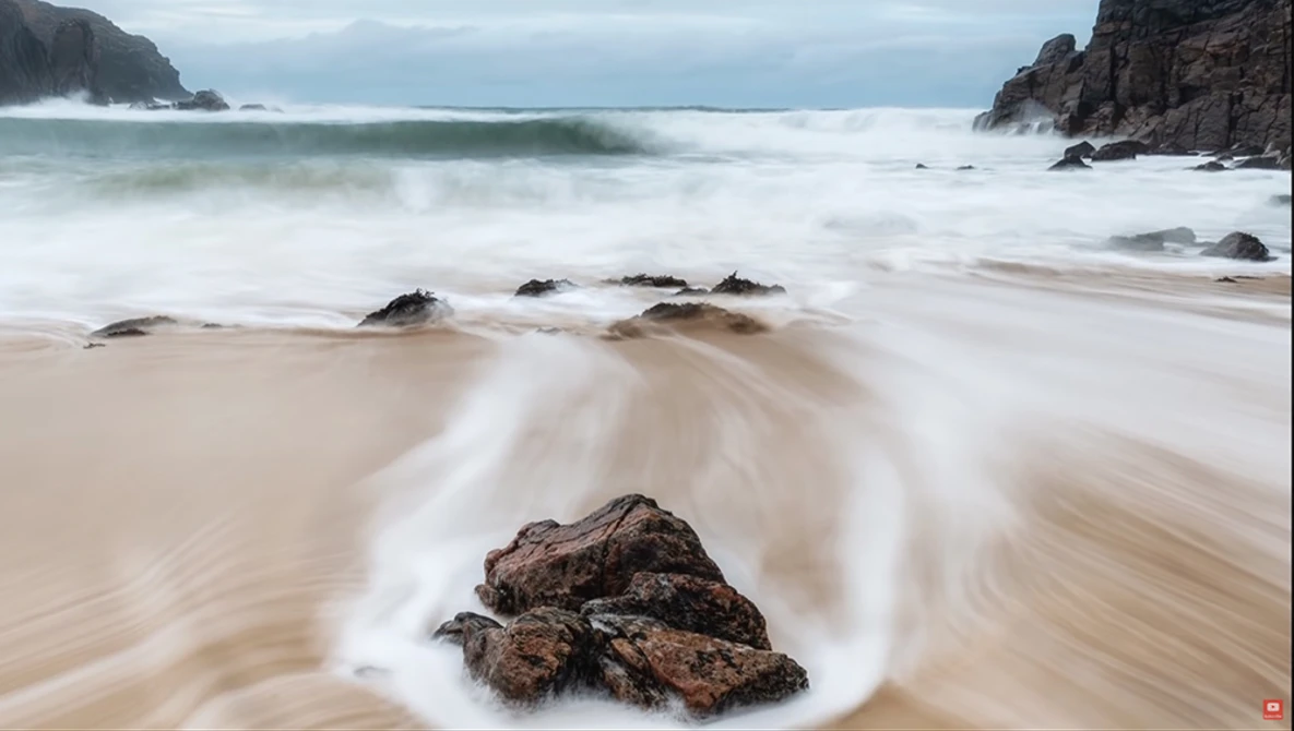 Long exposure seascape with swirling water around rocky outcrops on sandy beach.