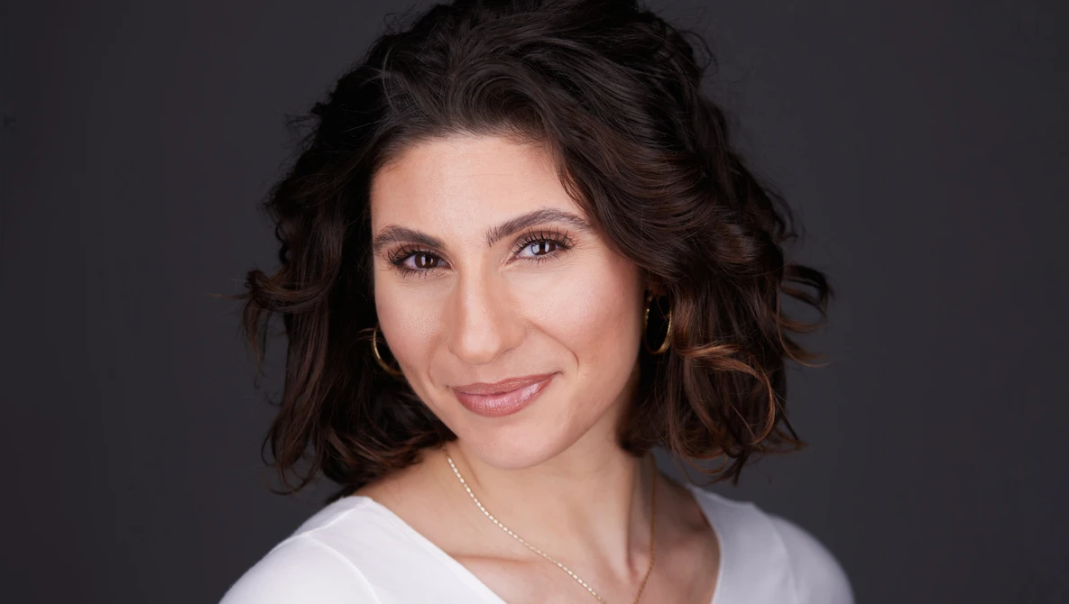 Woman with dark wavy shoulder-length hair smiling at camera against gray background.