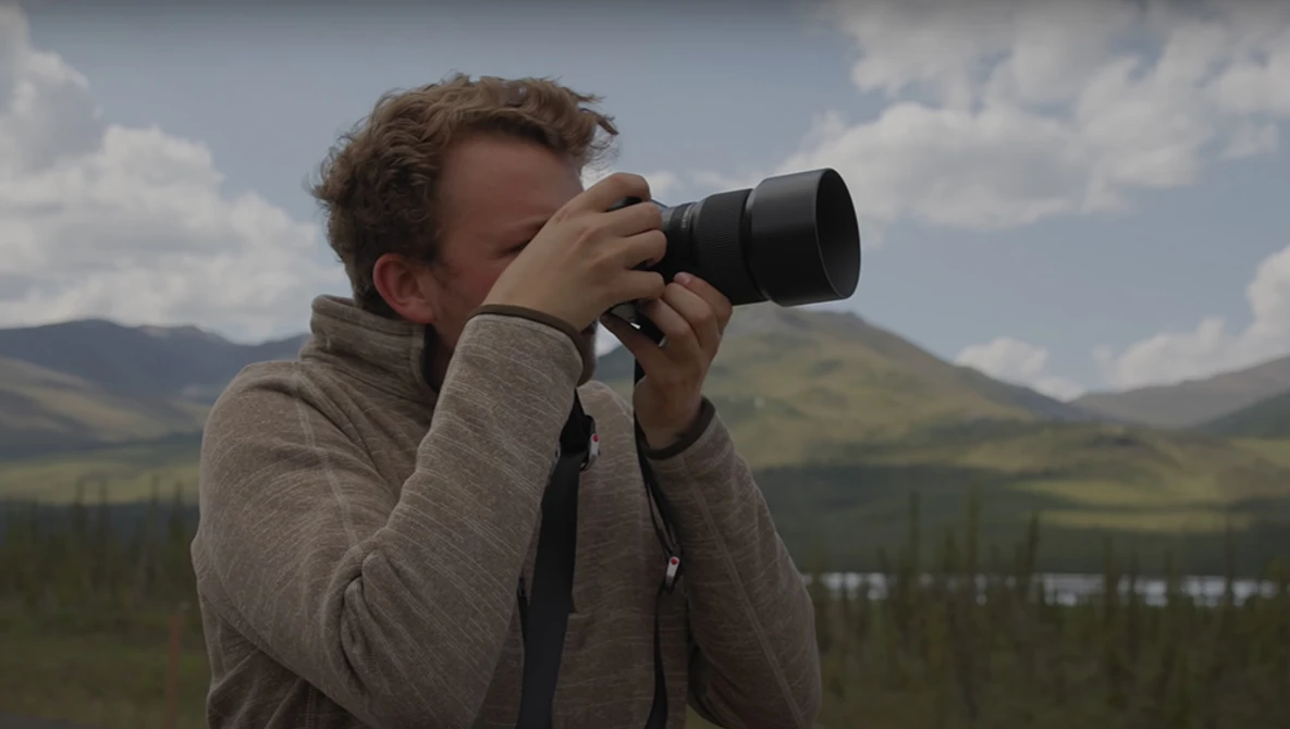 Photographer holding a telephoto lens up to their eye, photographing a mountainous landscape under partly cloudy skies.
