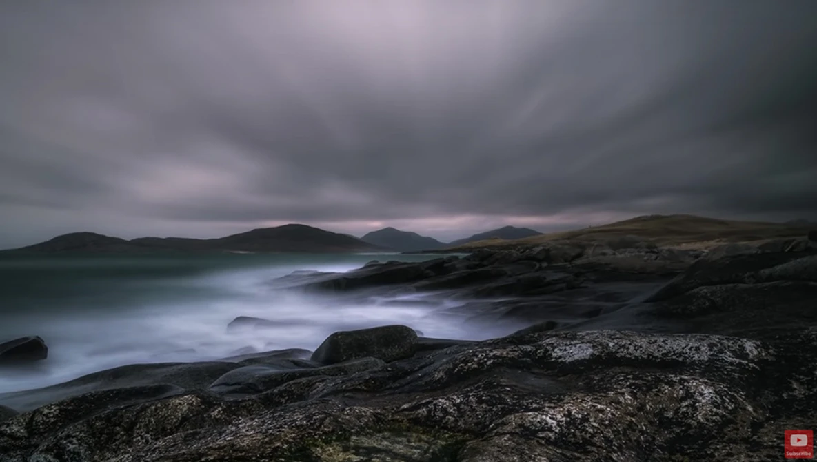 Rocky coastline with misty waves under stormy gray clouds and distant mountains.