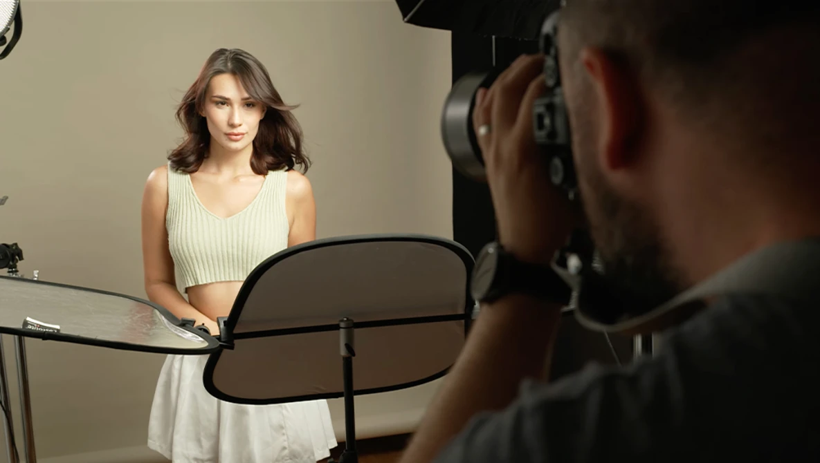 Photographer holding a camera on a reflector during a studio portrait session with a woman in a cream-colored dress.