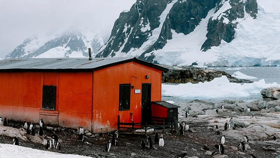 Bright red cabin with black windows on rocky shore, snow-capped mountains and glaciers behind it, penguins on ground nearby.