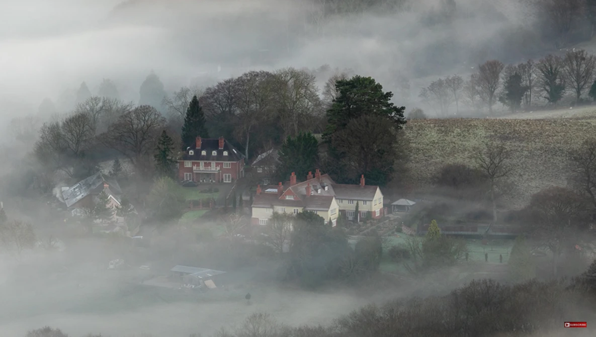 Medieval castle and village shrouded in thick morning mist among bare winter trees.