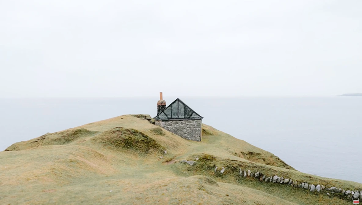 Solitary stone house with red chimney sits on barren hilltop under overcast sky.