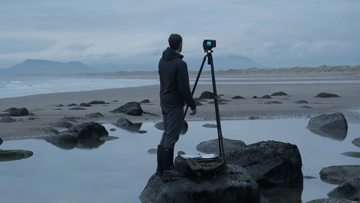 Photographer standing on rocks with camera mounted on tripod overlooking a rocky beach and ocean.