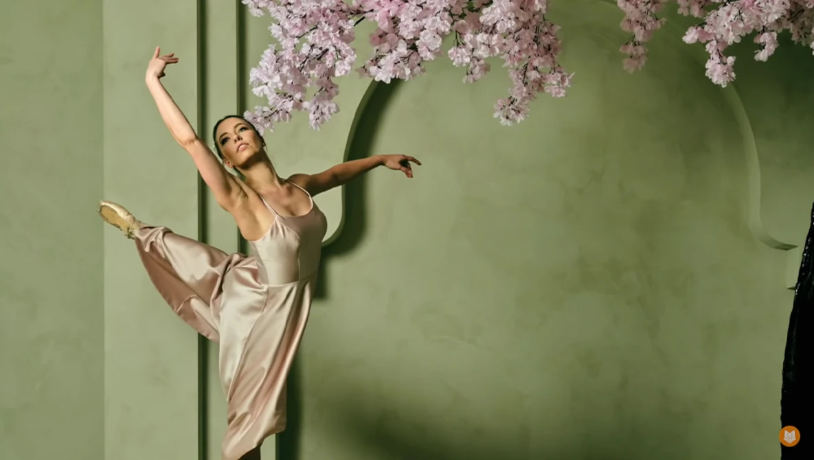 Ballet dancer in arabesque position wearing pale pink pointe shoes and satin dress against green wall with hanging lilac flowers.