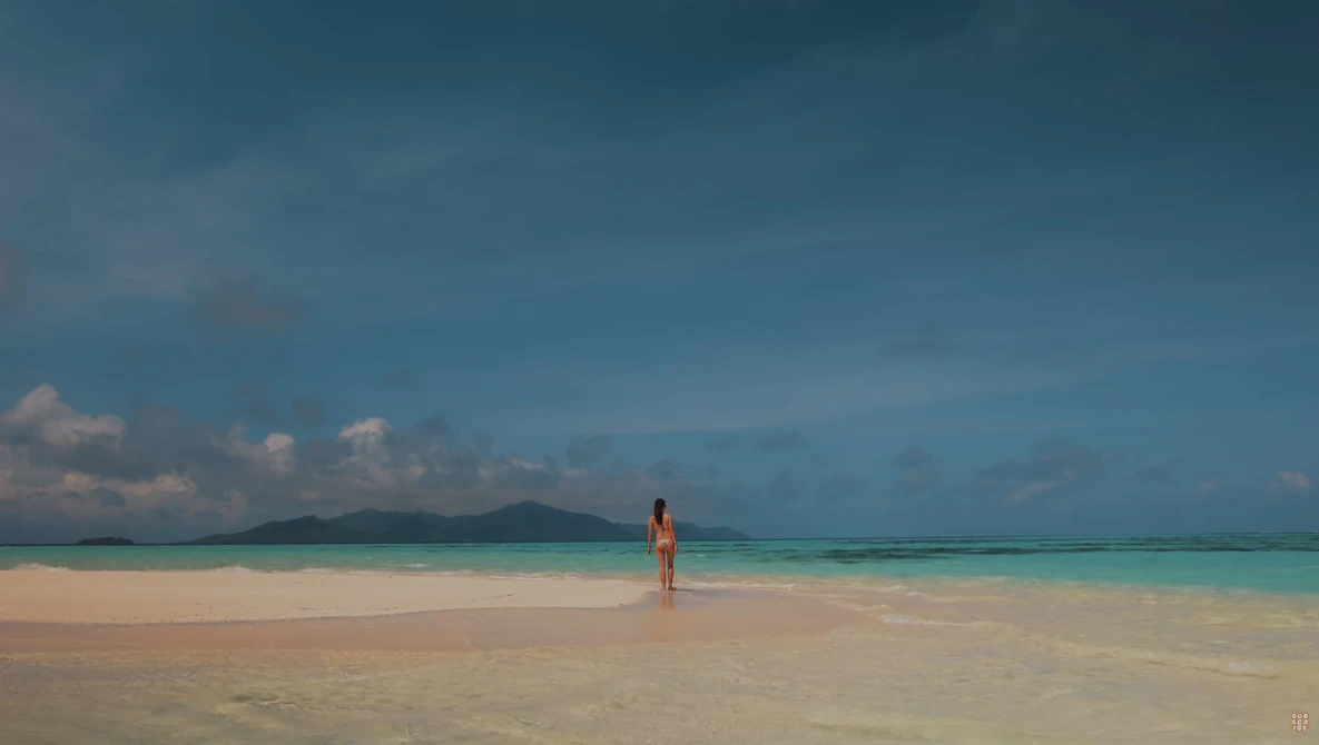 Solitary figure in orange standing in shallow turquoise water on a pristine beach with mountains visible across the water.