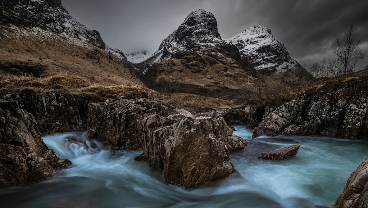 Snow-capped mountains frame a turquoise river flowing over dark rock formations.