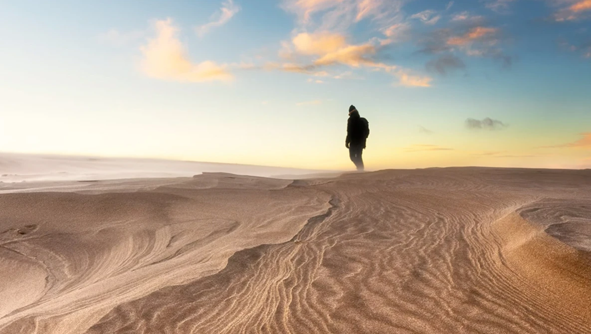 Silhouetted figure standing on rippled sand at sunset, with ocean and sky in background.