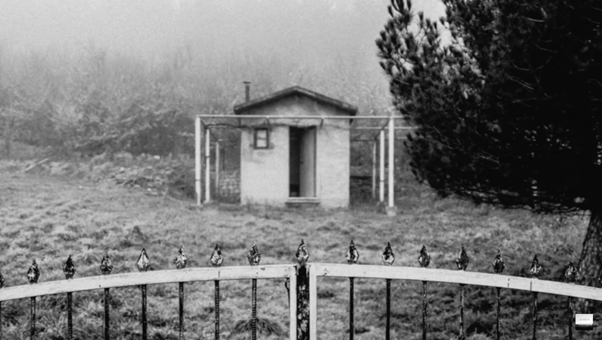 Small white wooden structure with open covered porch set in misty forest clearing behind metal fence.