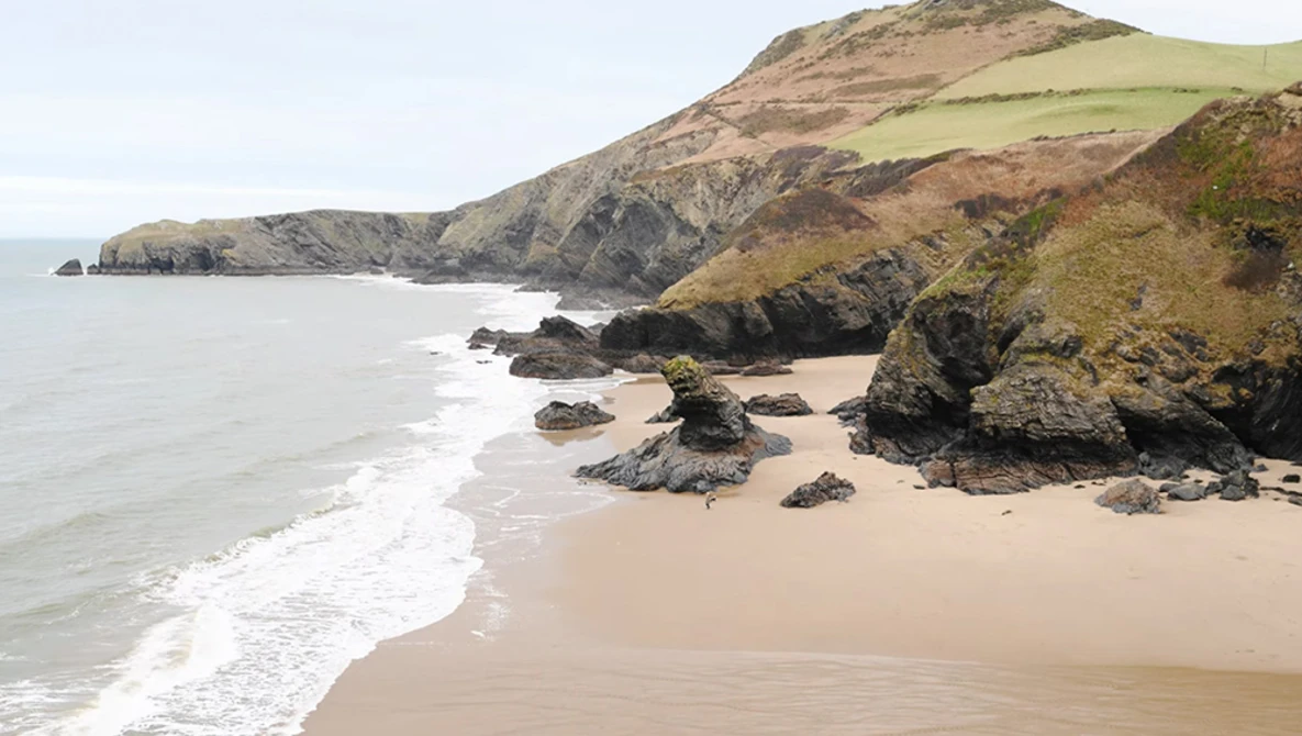 Rocky coastline with sandy beach, green hillsides, and breaking waves under overcast sky.