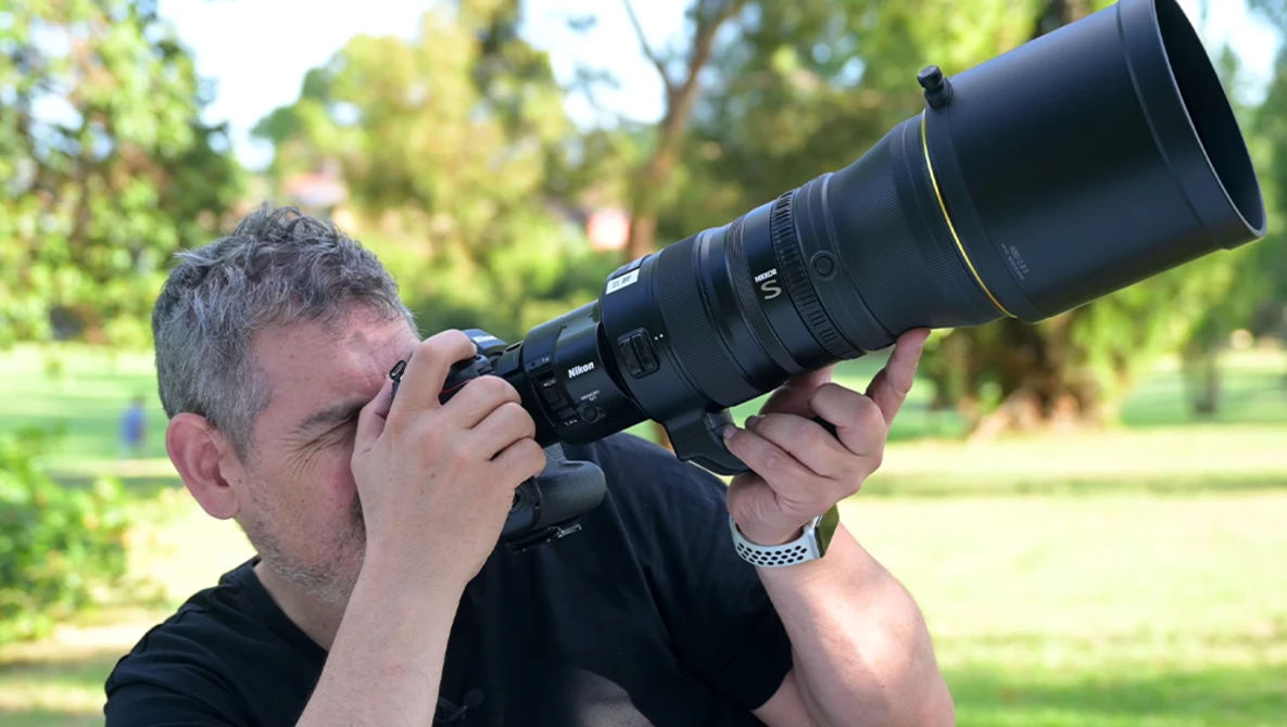 Photographer with gray hair holding a camera with a large telephoto lens to his eye outdoors.