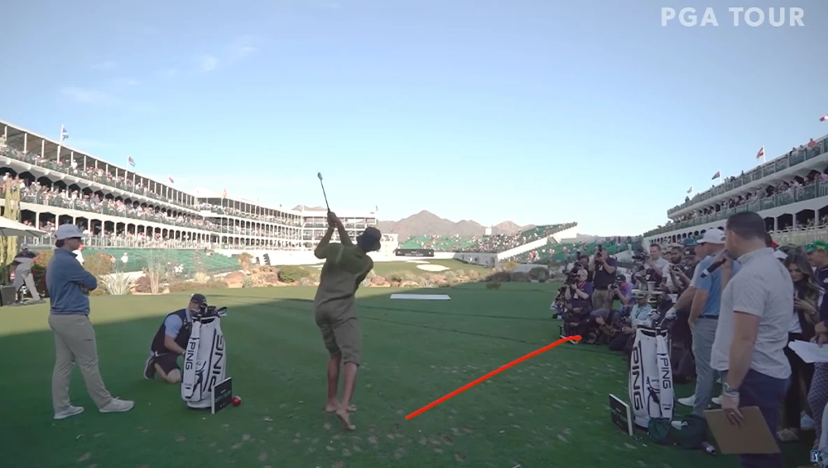 Professional golfer mid-swing on a tournament fairway with spectators and grandstands visible.
